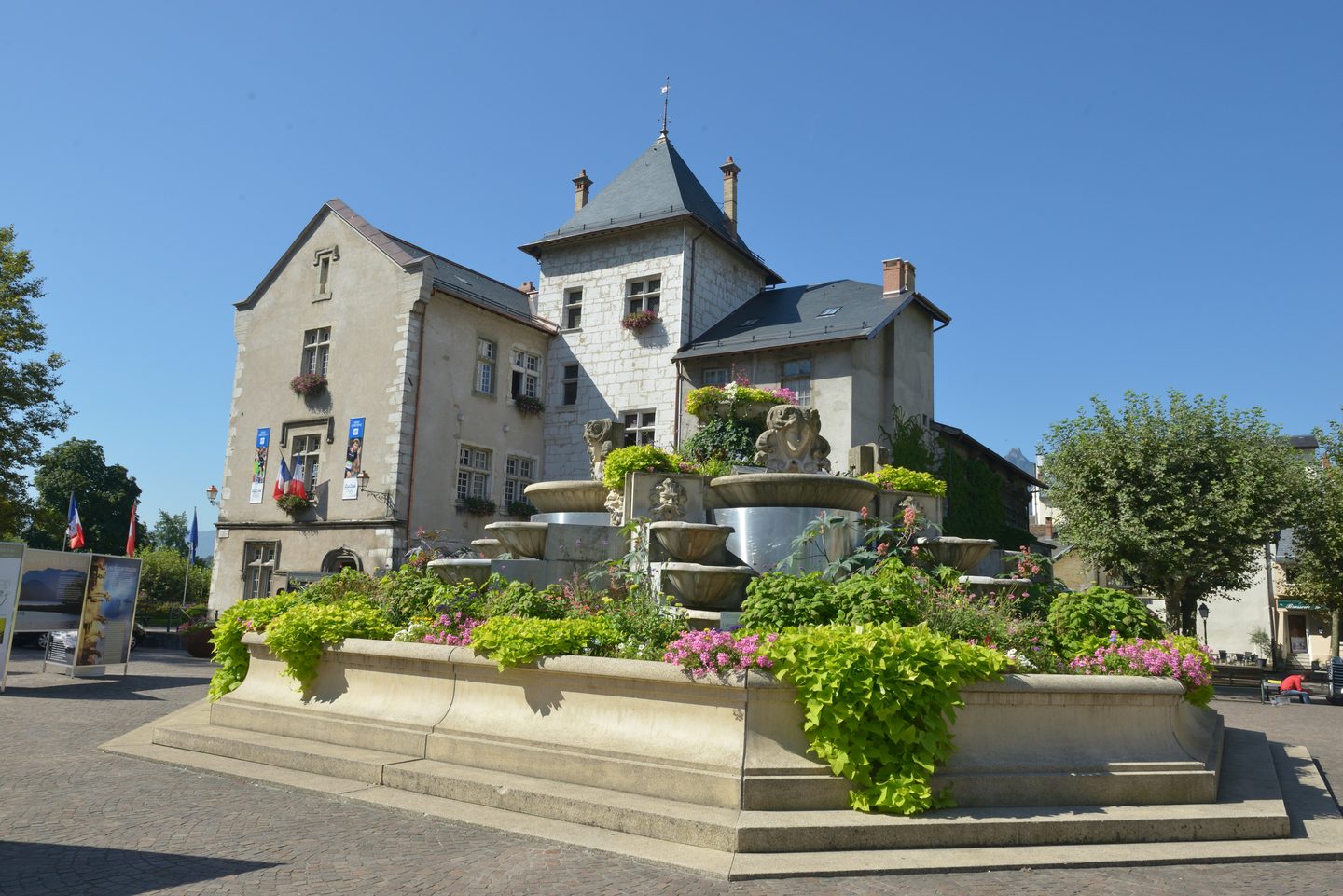 Bâtiment ancien à tourelle, orné de drapeaux, derrière une fontaine fleurie.