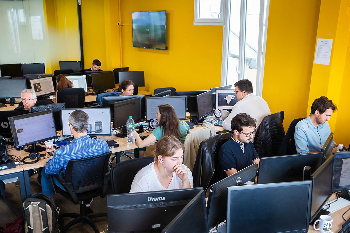La salle de rédaction de 6Médias, aux murs jaunes : une douzaine de journalistes travaillent à leurs écrans.