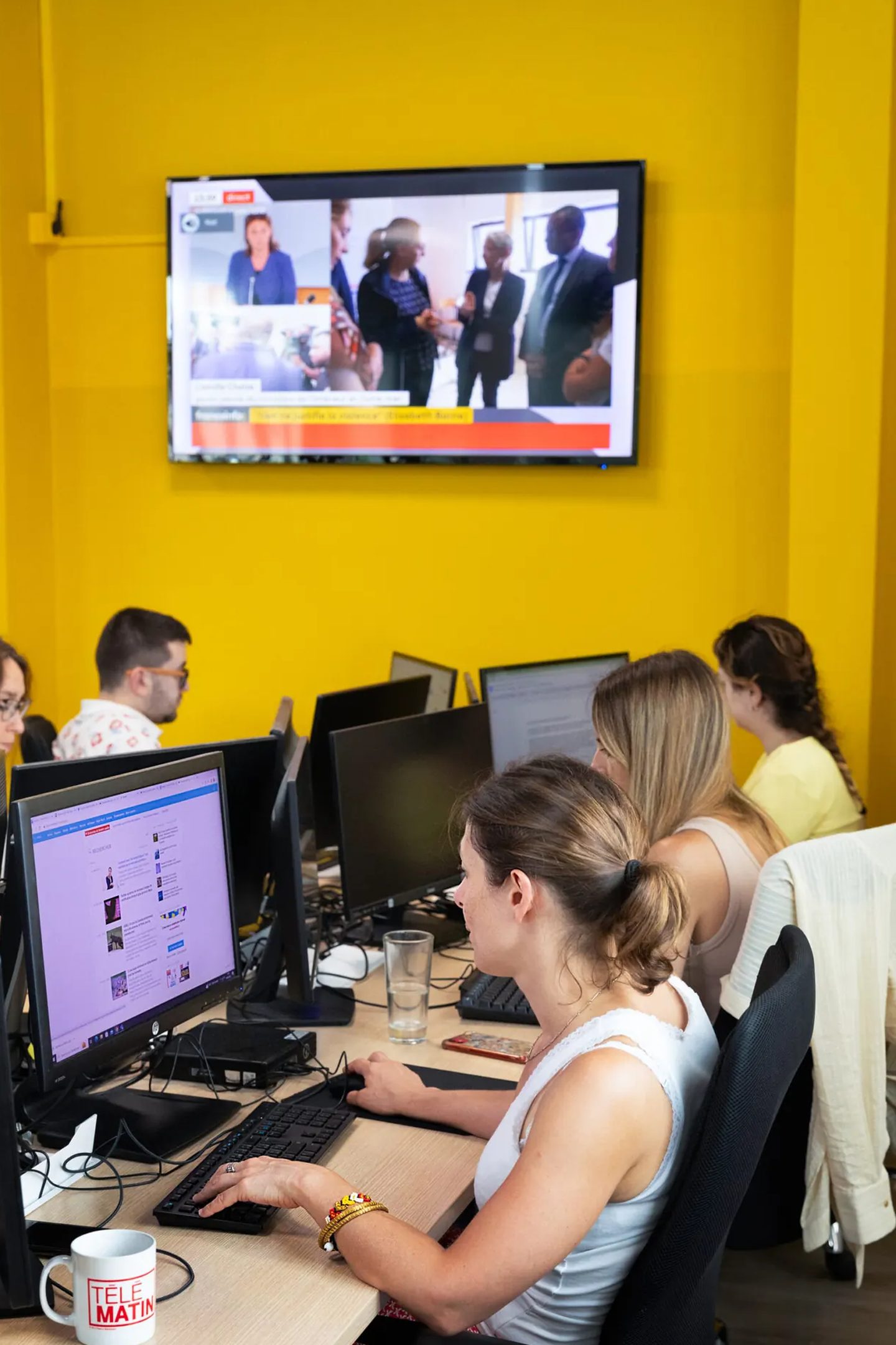 Des journalistes de 6Médias travaillent sur plusieurs écrans sous un téléviseur allumé sur une chaîne d'information, dans la salle de rédaction aux murs jaunes.